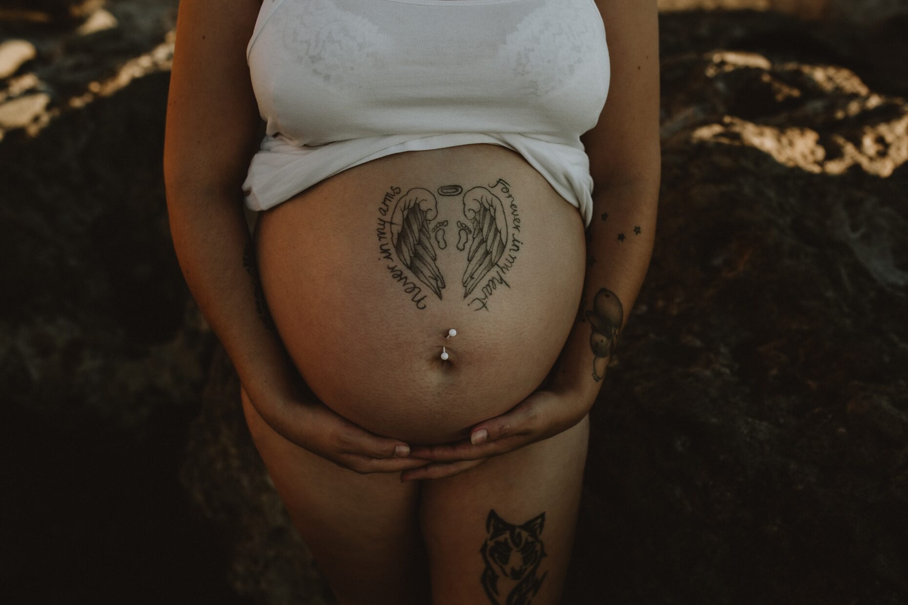 Mum holding belly on the beach, sun going down, tattoos about baby